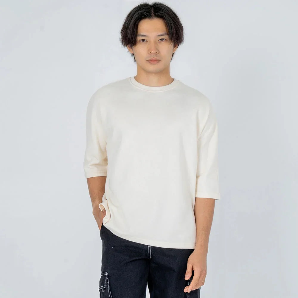 Hombre con camiseta beige de manga tres cuartos y pantalón oscuro, fondo neutro