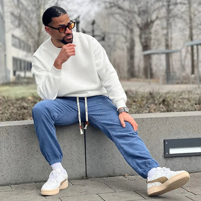 Hombre con sudadera blanca, pantalones jogger azul y zapatillas blancas, sentado al aire libre.